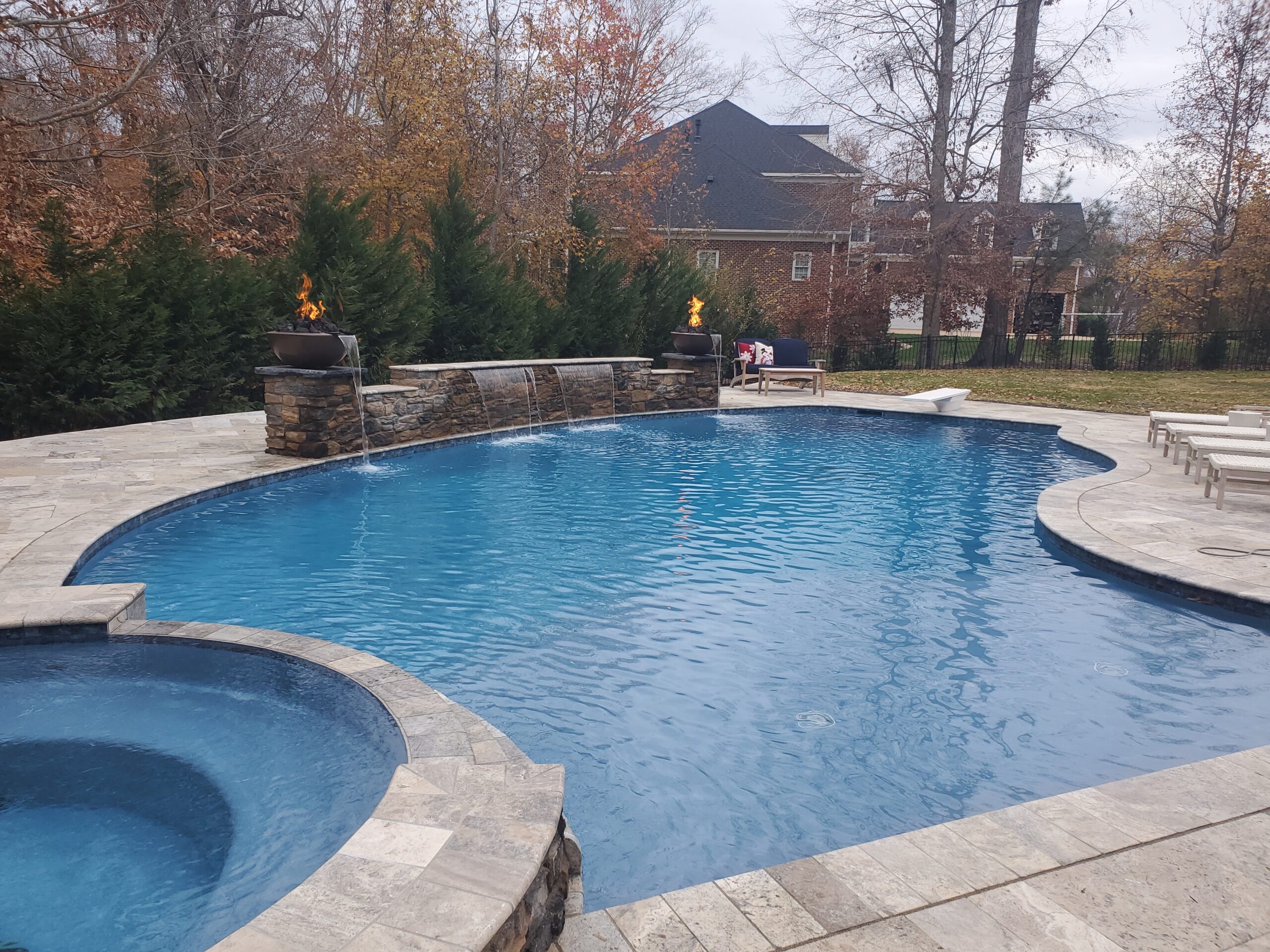 Freeform gunite pool with raised spa, stone waterfall wall, and fire bowls on a stone pool deck in a backyard.