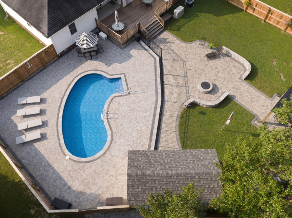 Drone photo of a freeform vinyl liner inground pool with gray paver deck, lounge chairs, and backyard patio layout.