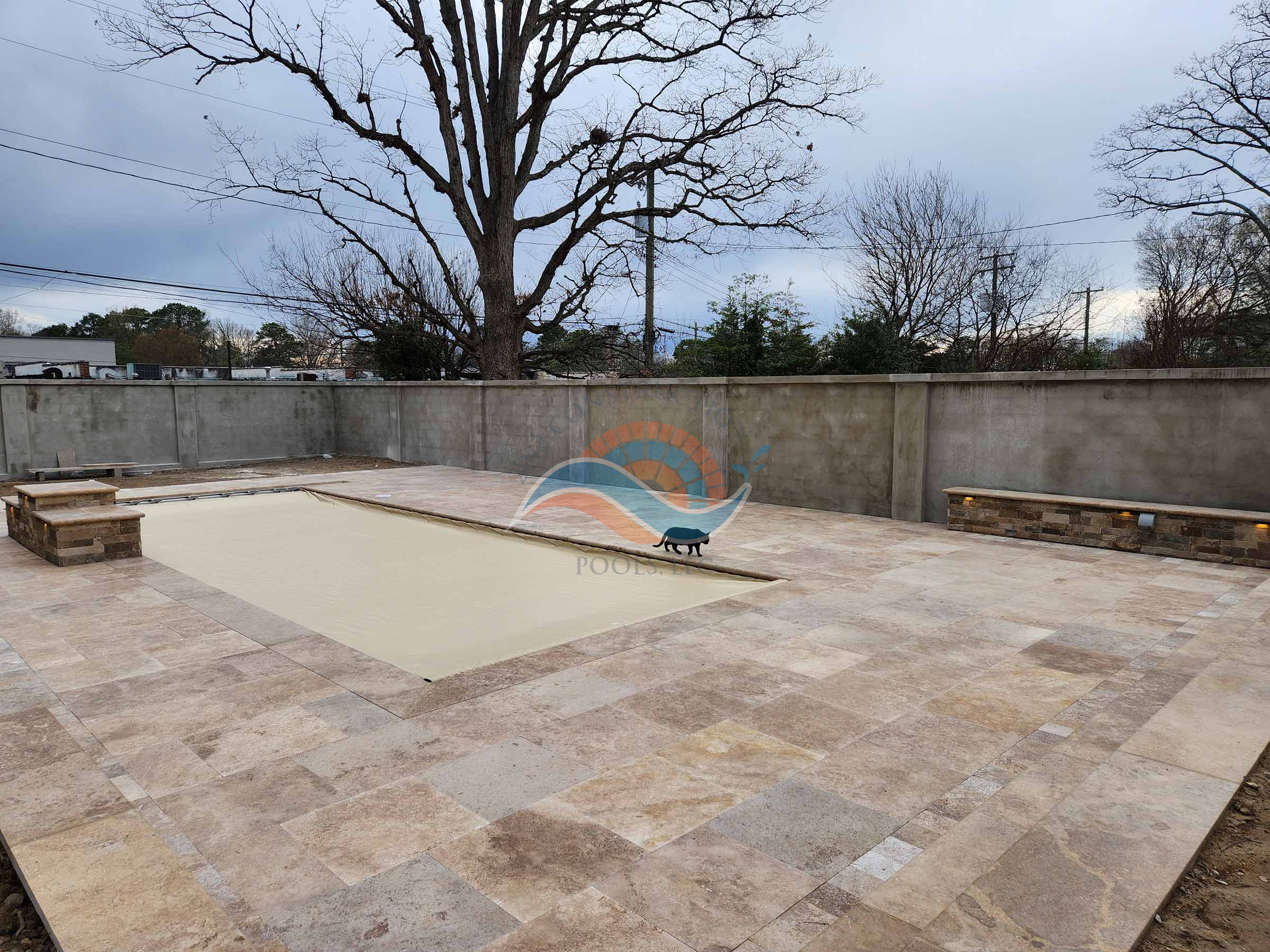 Fiberglass inground pool with automatic safety cover closed, large stone patio, and tall privacy wall in a backyard.