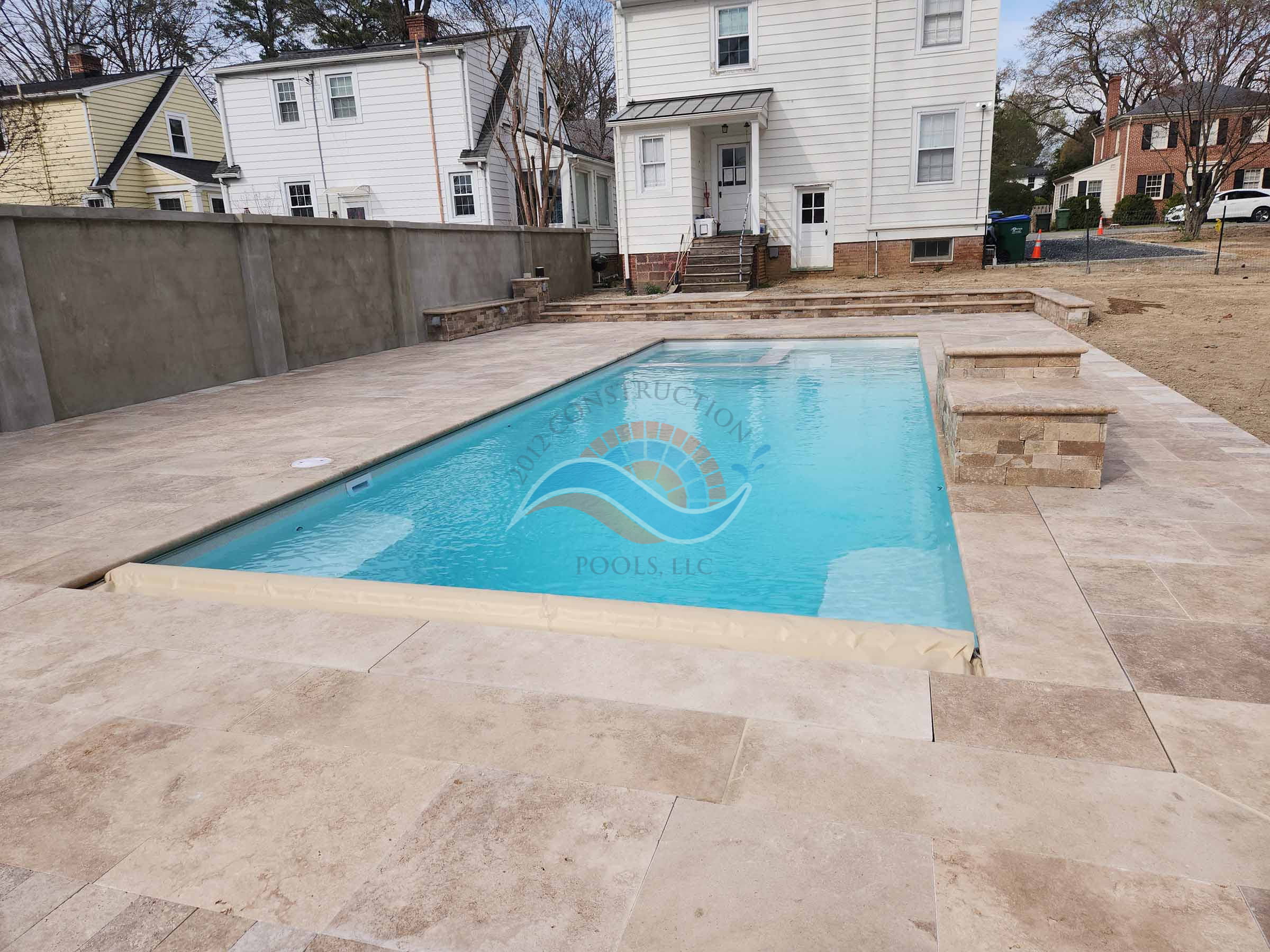 Fiberglass inground swimming pool with stone patio, built-in bench seating, and tall privacy wall in a residential backyard.
