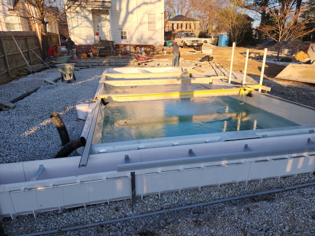 Fiberglass pool shell set in place with bracing over water, surrounded by gravel base during installation at a residential site.