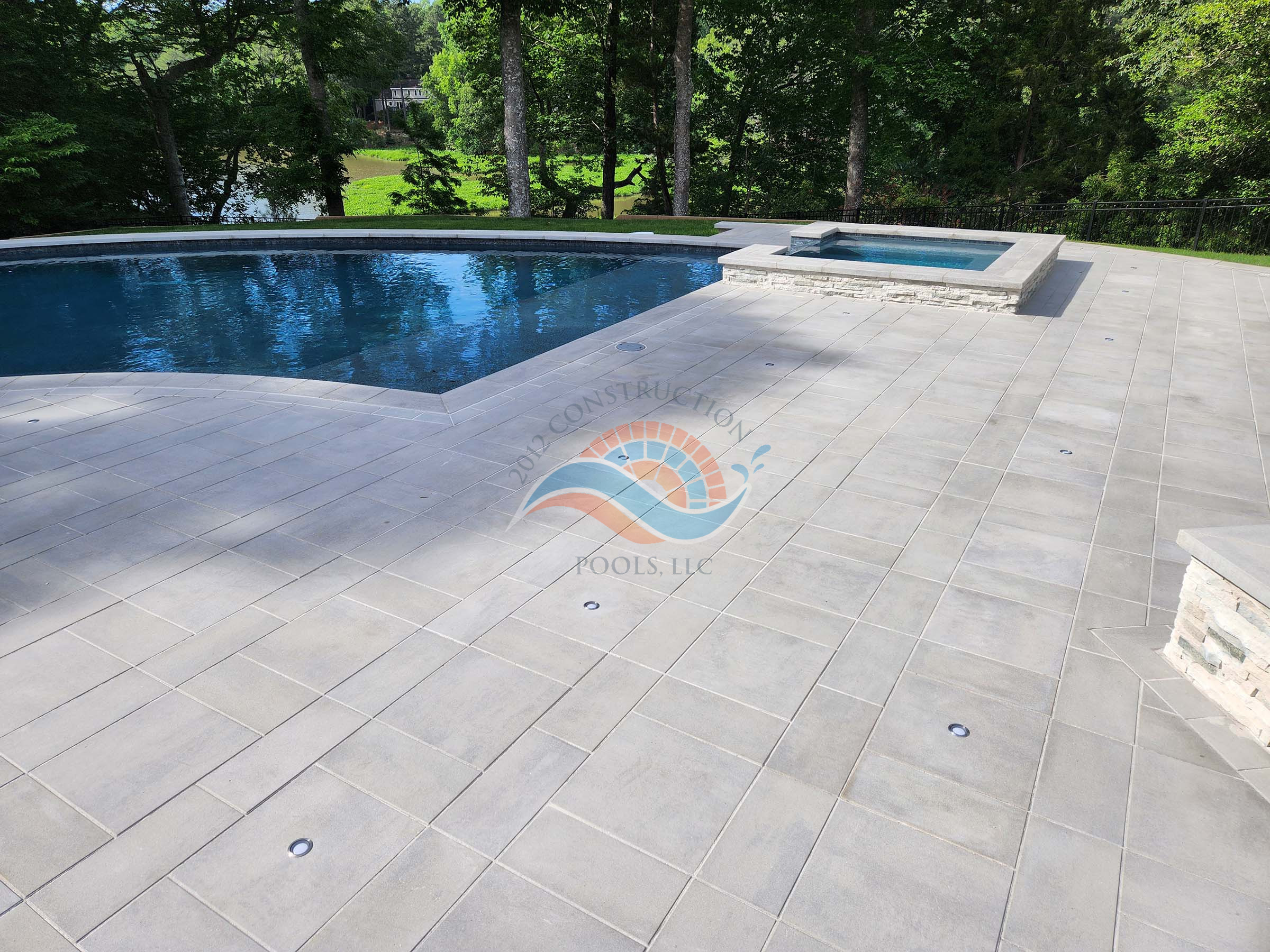 Finished gunite swimming pool with raised spa, stone veneer, and large paver pool deck in a wooded backyard.