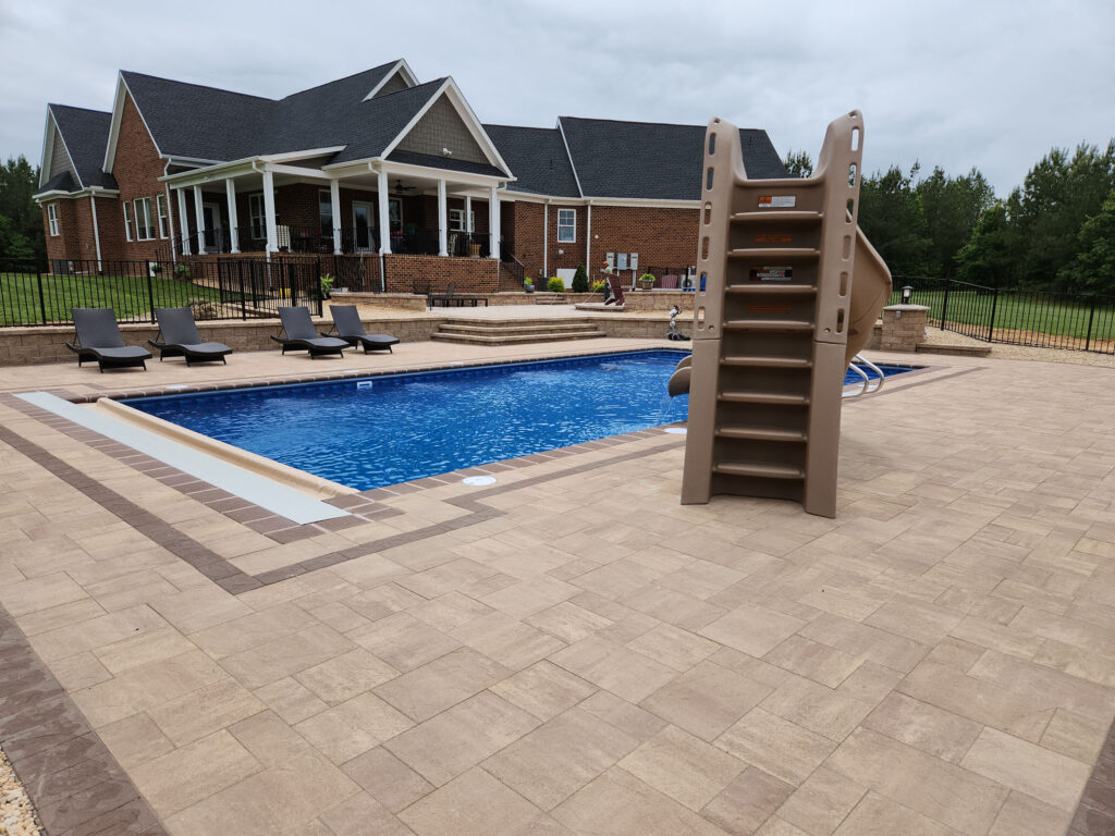 Finished vinyl liner inground swimming pool with large paver deck and pool slide in a residential backyard.