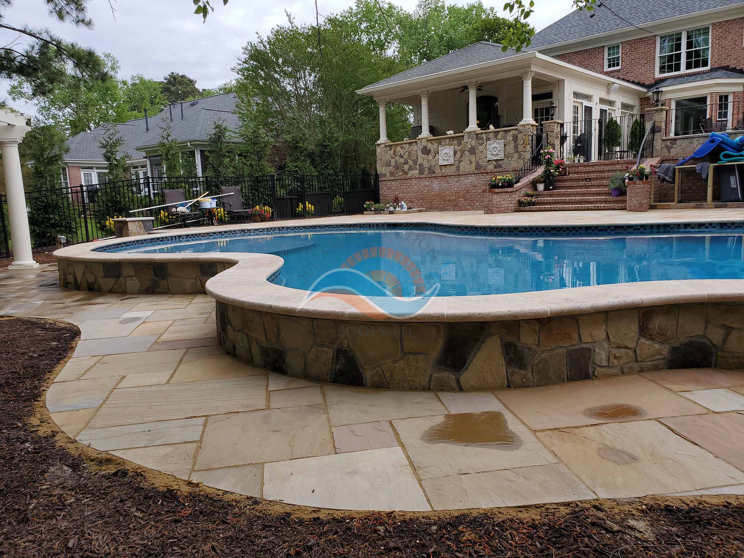 Freeform gunite pool with raised stone bond beam, natural stone coping, and flagstone deck behind a brick home.