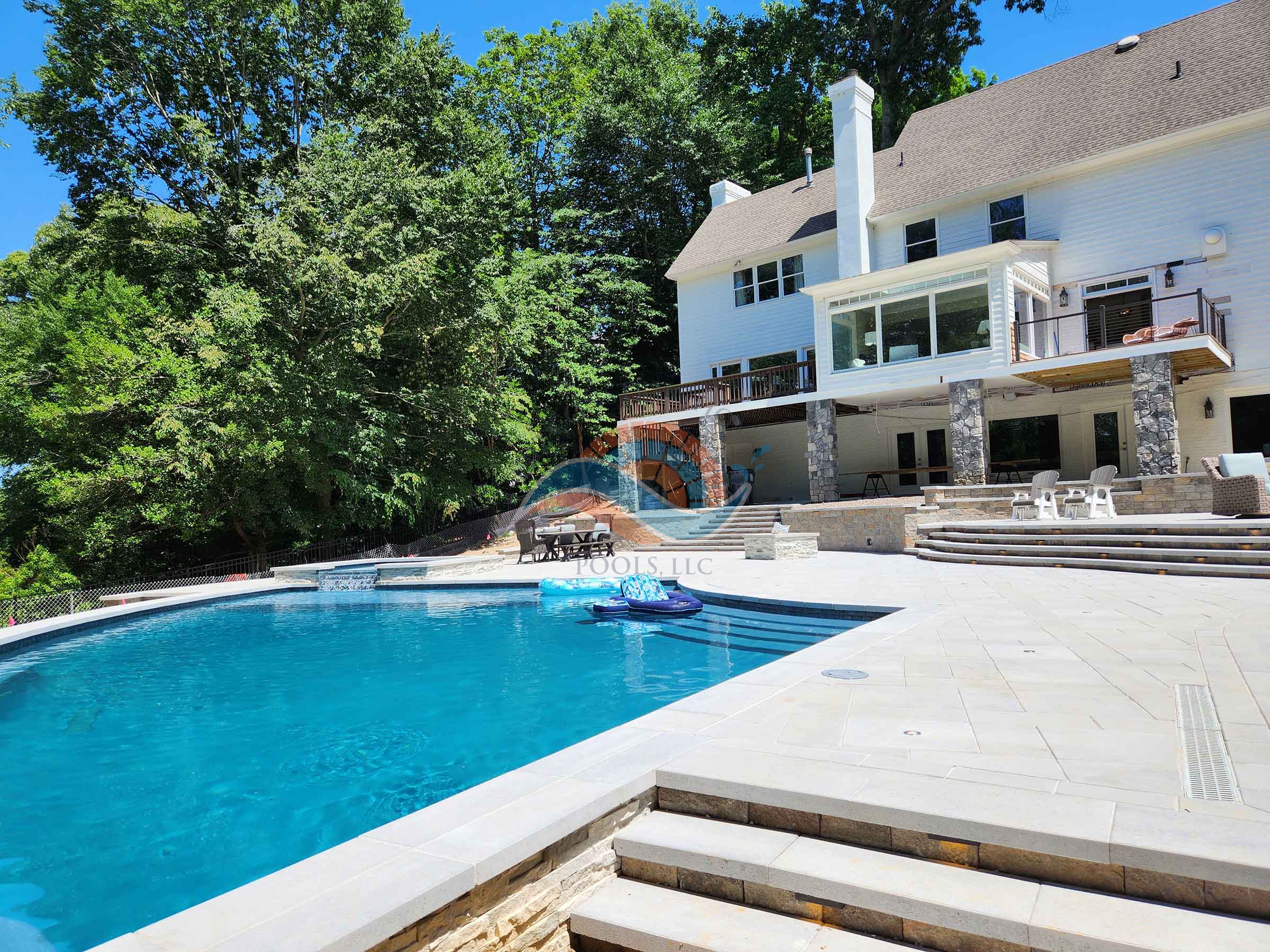 Gunite swimming pool with large light-colored patio, retaining walls, and tiered steps behind a white home on a wooded lot.