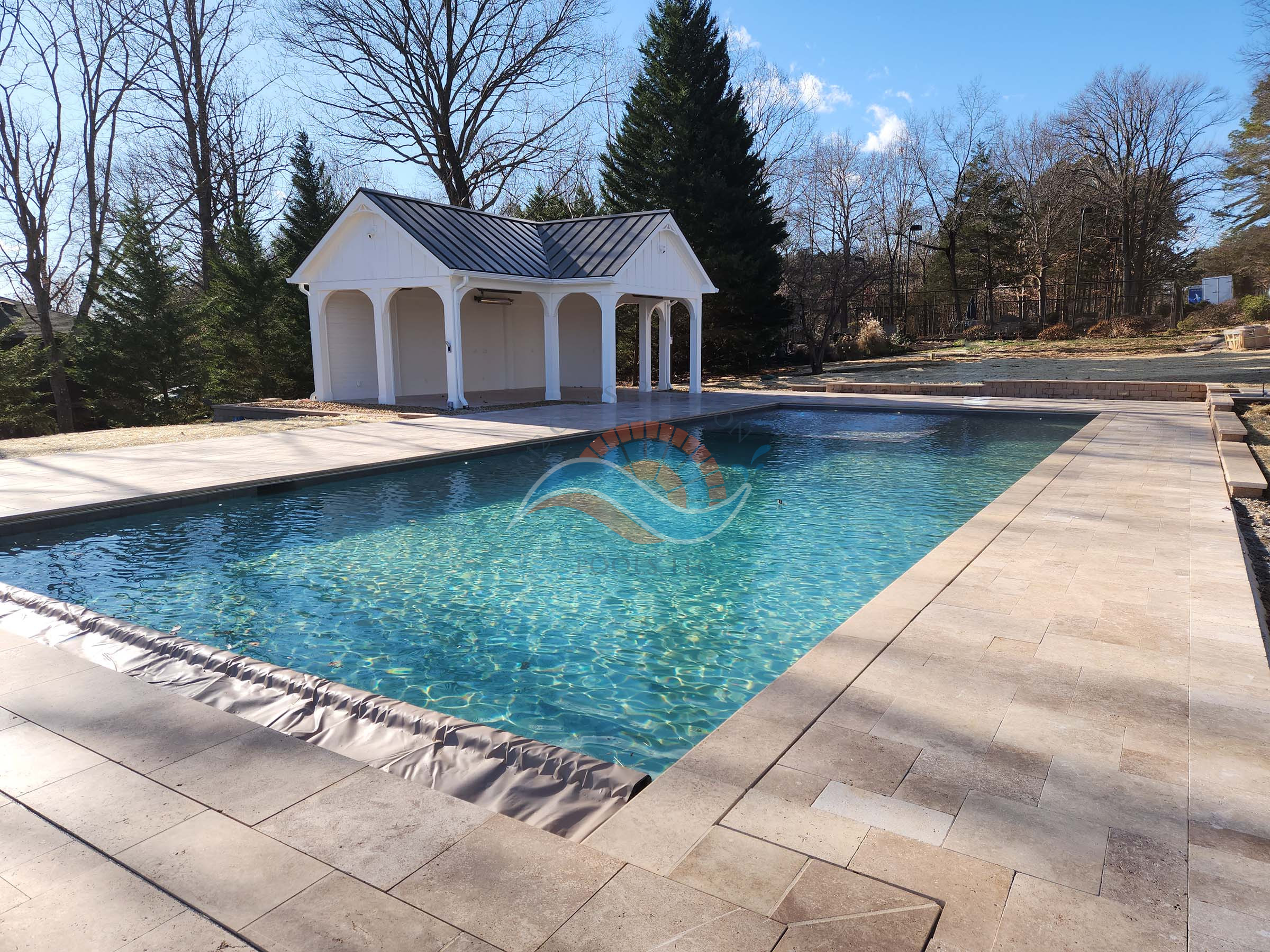 Rectangular in-ground pool with clear water, light stone paver deck, and a white pavilion with a dark metal roof in a landscaped backyard.